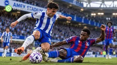 Um jogador do Brighton, com camisa listrada de azul e branco, salta para evitar uma entrada deslizante de um zagueiro do Crystal Palace, vestido de vermelho e azul, no Estádio Amex.
